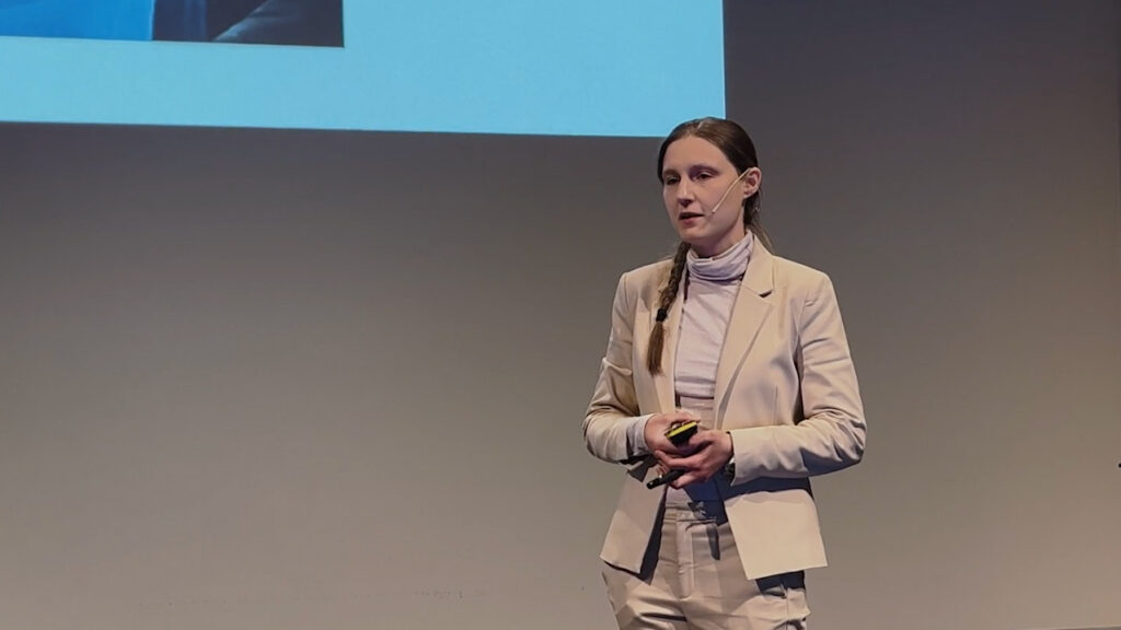 A woman in a light suit with a headset microphone stands on stage, holding a small device, giving a presentation. A projection screen is partially visible behind her.