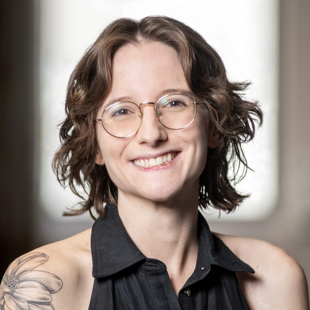 A person with short wavy brown hair, round glasses, and a flower tattoo on their shoulder, smiling and wearing a sleeveless black top, sits in front of a softly blurred indoor background.