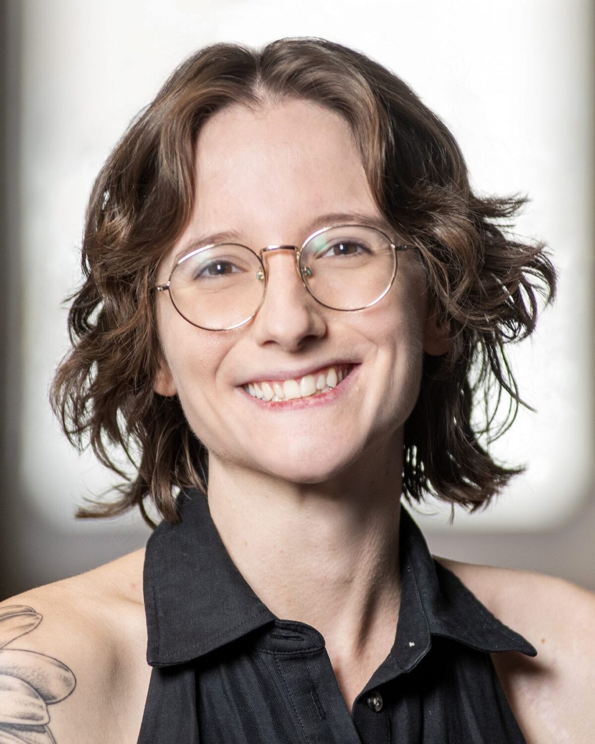 A person with short wavy brown hair, round glasses, and a flower tattoo on their shoulder, smiling and wearing a sleeveless black top, sits in front of a softly blurred indoor background.