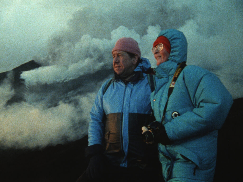 Two people in blue winter jackets and hats stand close together, one holding a camera, with smoke rising from a volcano in the background.