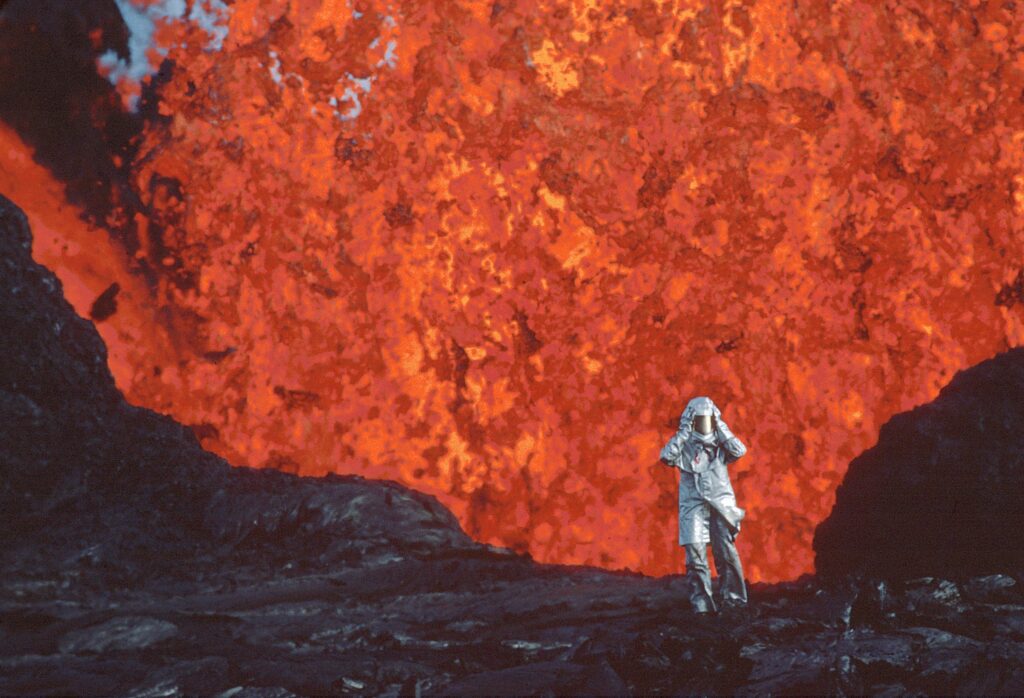A person in a silver heat-protective suit stands on dark volcanic rock in front of a large, bright orange lava explosion.