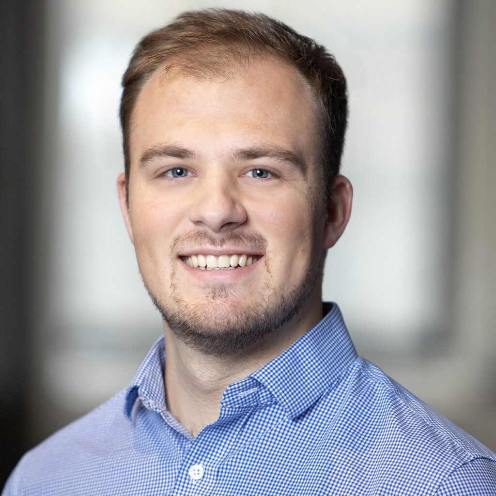 A young man with short light brown hair and a trimmed beard smiles at the camera, wearing a blue checkered button-down shirt. The background is softly blurred in neutral tones.
