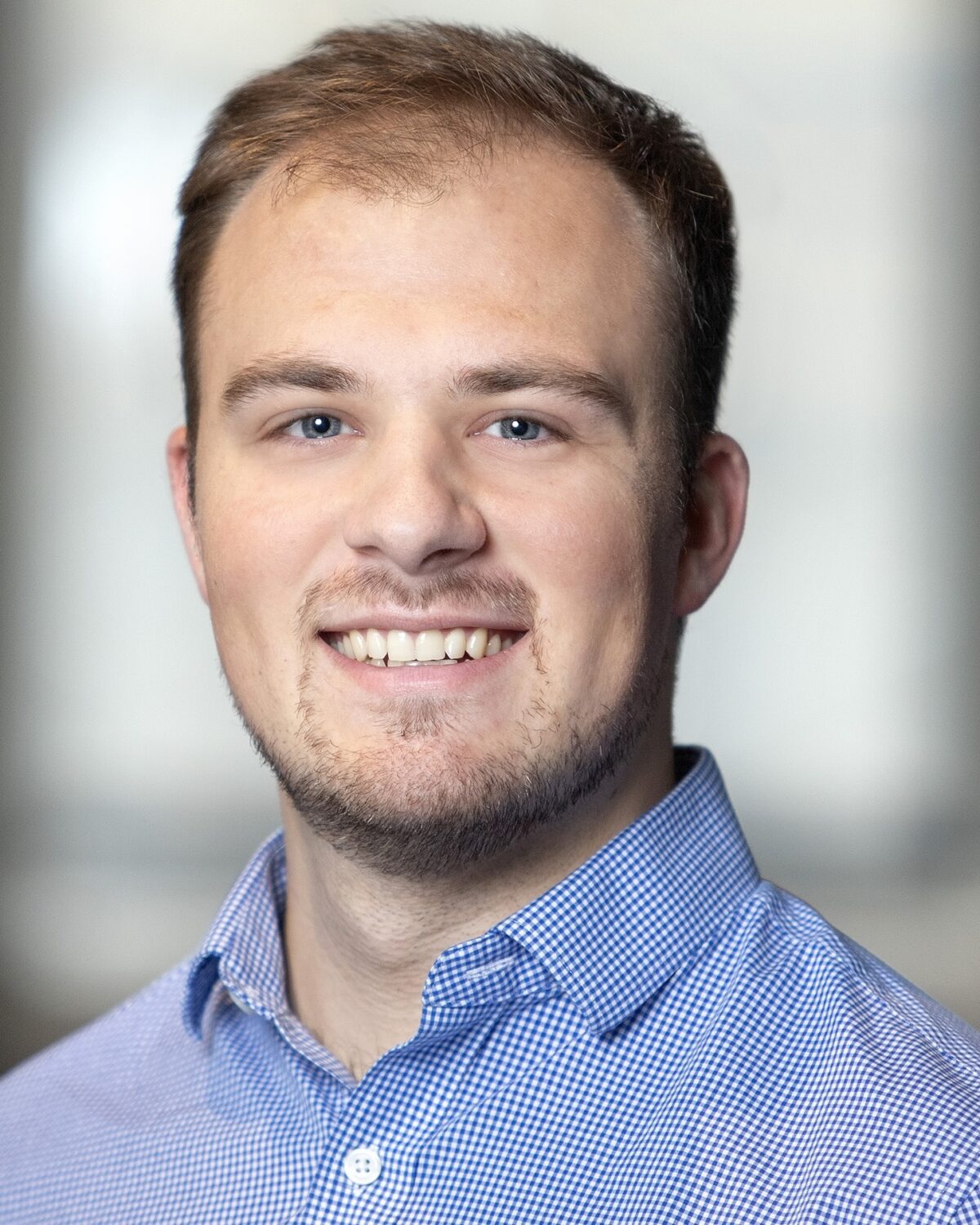 A young man with short light brown hair and a trimmed beard smiles at the camera, wearing a blue checkered button-down shirt. The background is softly blurred in neutral tones.