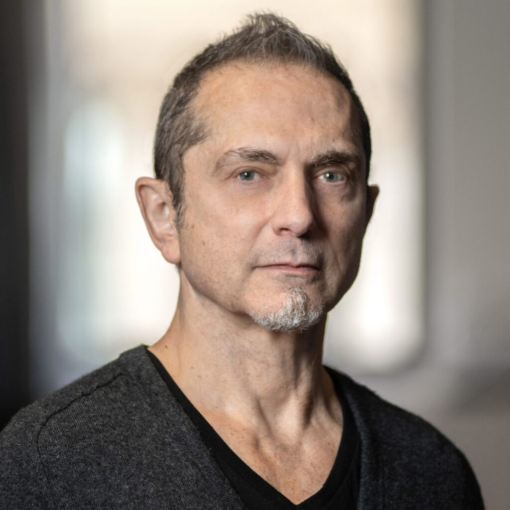 A middle-aged man with short hair and a small goatee looks at the camera. He wears a dark V-neck shirt and stands indoors with blurred windows in the background.
