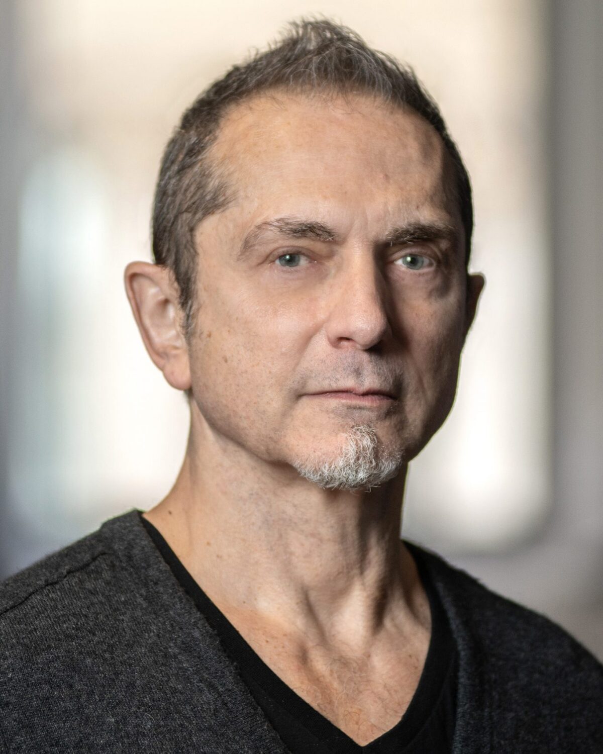 A middle-aged man with short hair and a small goatee looks at the camera. He wears a dark V-neck shirt and stands indoors with blurred windows in the background.
