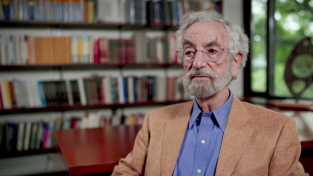 An older man with gray hair and a beard, wearing glasses, a tan jacket, and blue shirt sits in front of bookshelves filled with books.