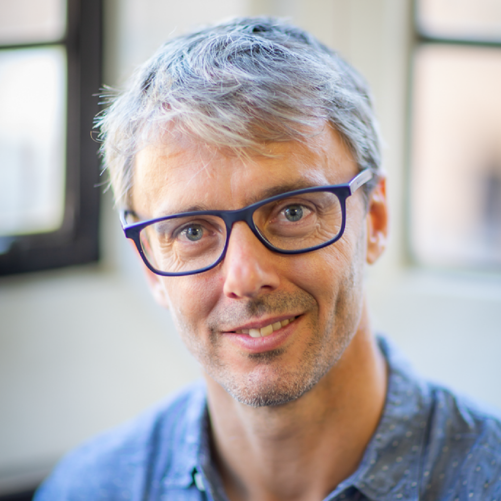A middle-aged man with gray hair, wearing dark-rimmed glasses and a blue patterned shirt, smiles at the camera in a softly lit room with blurred windows in the background.