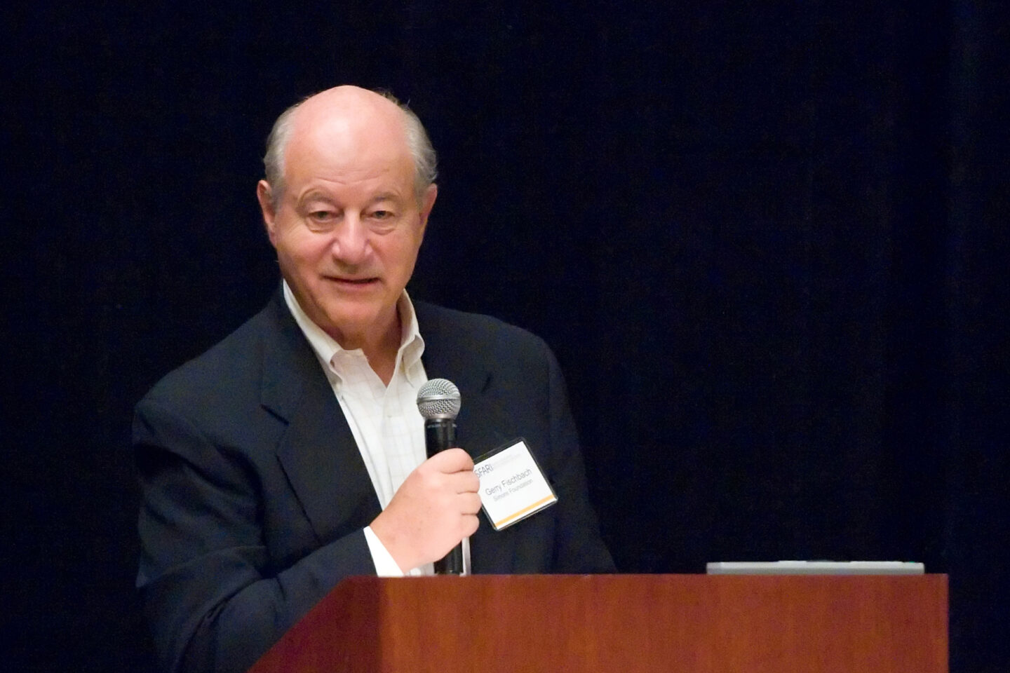 An older man in a suit speaks into a microphone at a podium, wearing a name tag, with a dark background.