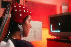 A person wearing a red EEG cap looks at a computer screen displaying a simple video game in a red-lit room.