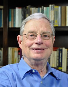 An older man with glasses and gray hair, wearing a blue shirt, smiles while sitting in front of a bookshelf filled with books.
