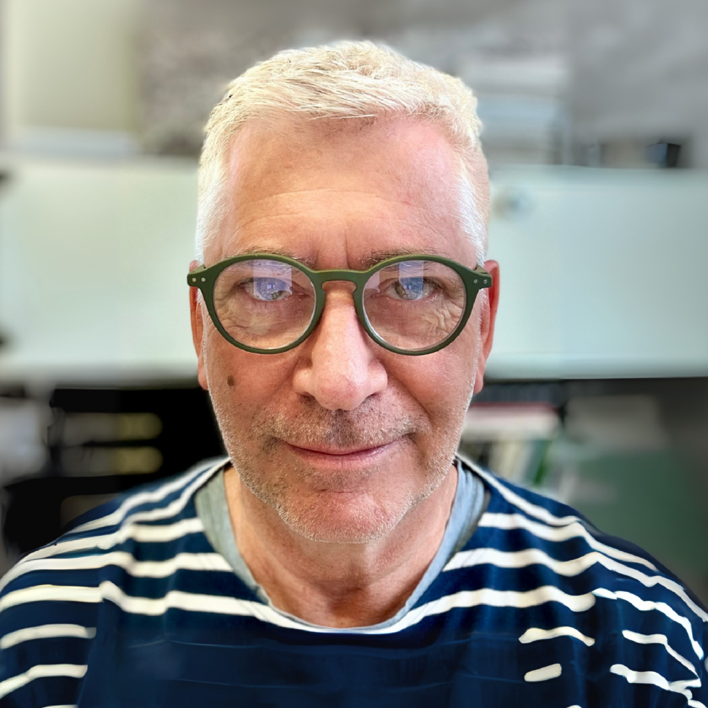 Older man with short gray hair and green round glasses, wearing a navy and white striped shirt, smiles slightly in a softly blurred indoor office setting.