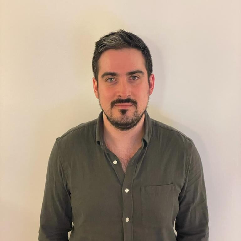 A man with short dark hair, light skin, and a trimmed beard stands against a plain off-white wall, wearing a dark green button-up shirt with the top buttons undone.