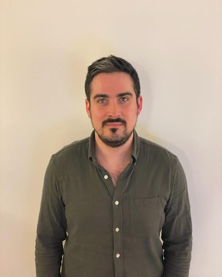 A man with short dark hair, light skin, and a trimmed beard stands against a plain off-white wall, wearing a dark green button-up shirt with the top buttons undone.
