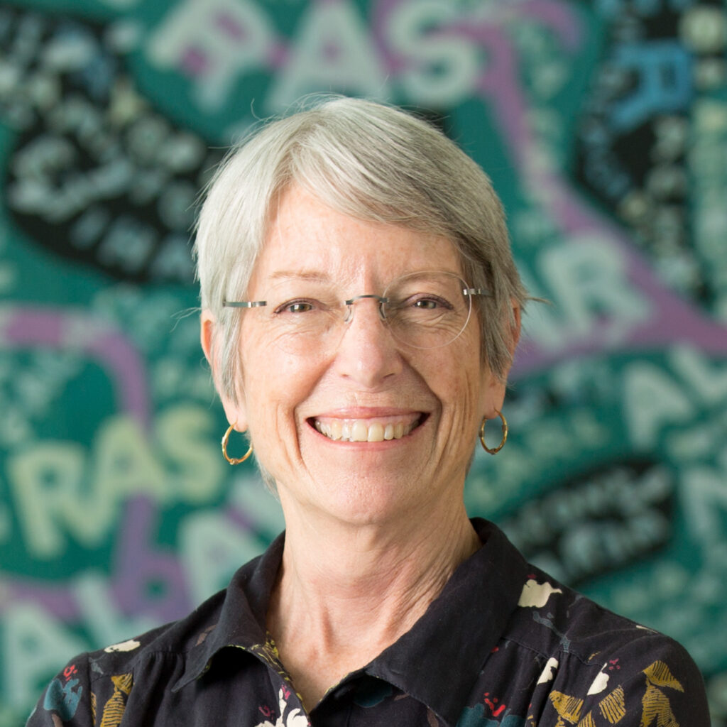 An older woman with short gray hair and glasses smiles at the camera. She is wearing hoop earrings and a dark patterned shirt. The background is colorful and abstract with the word RAISE repeatedly visible.