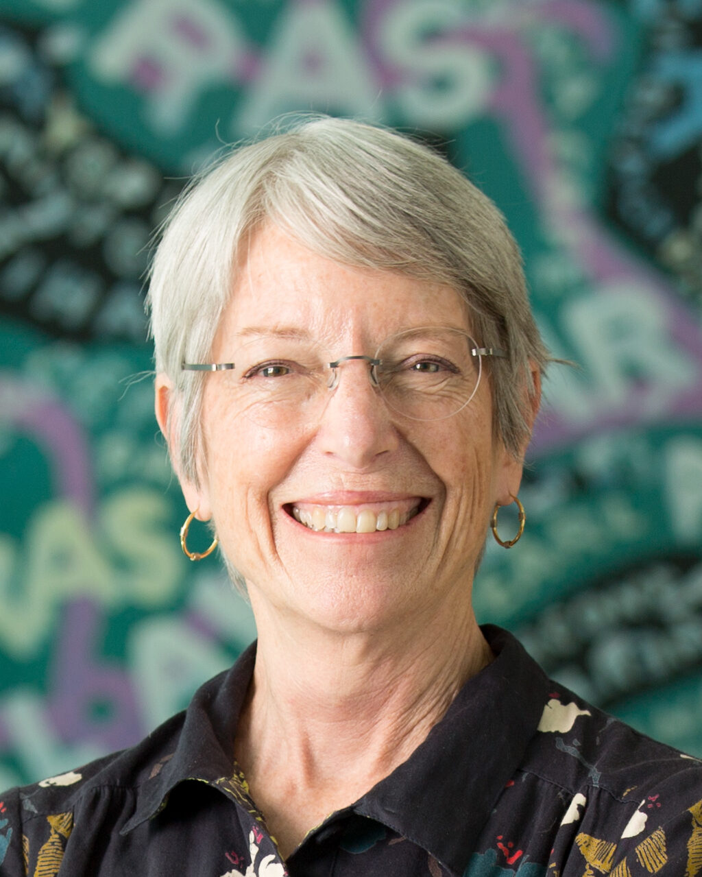 An older woman with short gray hair and glasses smiles at the camera. She is wearing hoop earrings and a dark patterned shirt. The background is colorful and abstract with the word RAISE repeatedly visible.