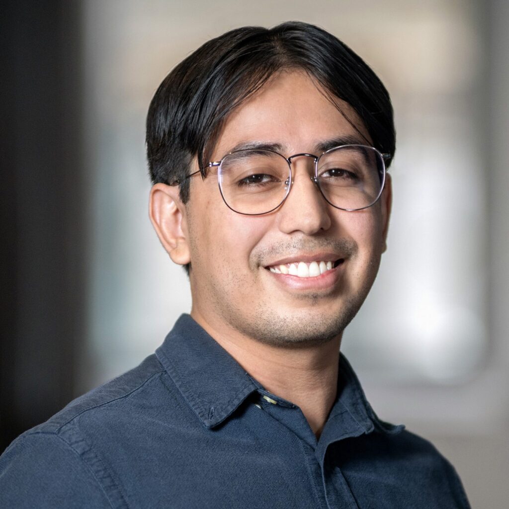 A person with short dark hair and glasses smiles at the camera, wearing a dark button-up shirt. The background is softly blurred.