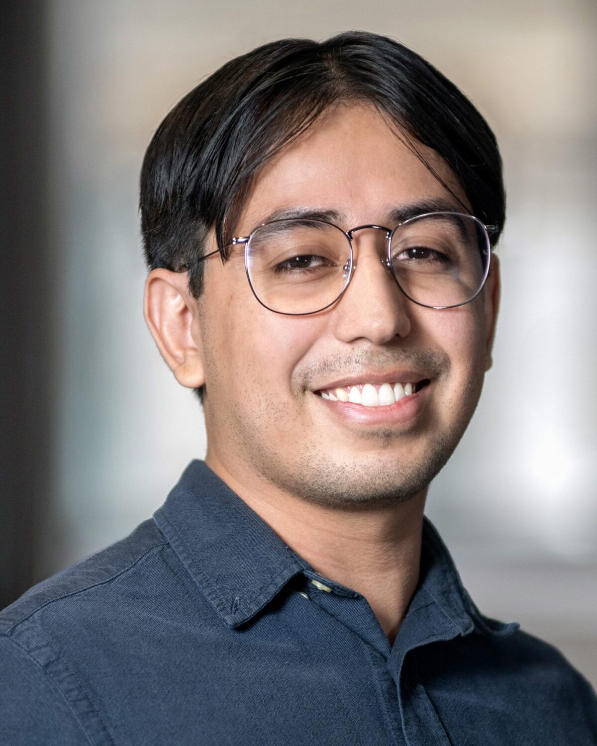 A person with short dark hair and glasses smiles at the camera, wearing a dark button-up shirt. The background is softly blurred.