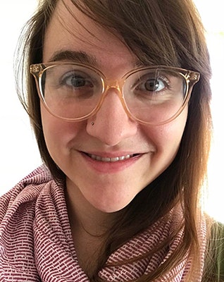 A person with light skin, long brown hair, glasses, and a nose piercing smiles at the camera, wearing a pink and white patterned scarf.