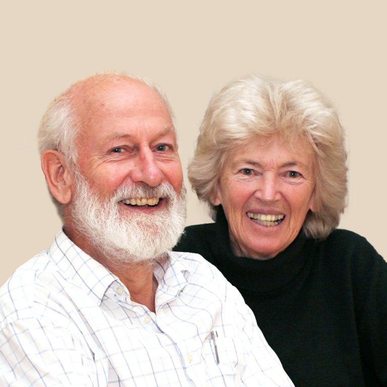An older man with a white beard and a woman with light hair are smiling, sitting close together against a plain beige background.