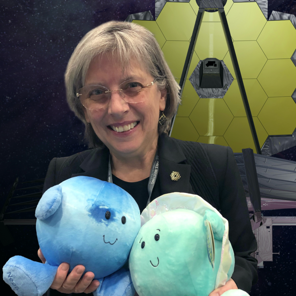A smiling woman with glasses holds two blue plush toys, standing in front of an image of the James Webb Space Telescope with its gold hexagonal mirrors visible.