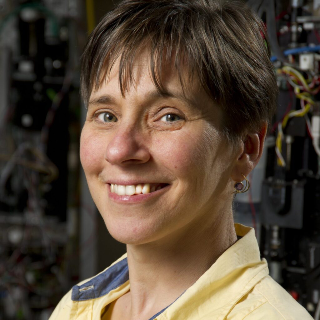 A person with short brown hair, wearing a yellow collared shirt, is smiling at the camera. The background shows blurred machinery and cables.