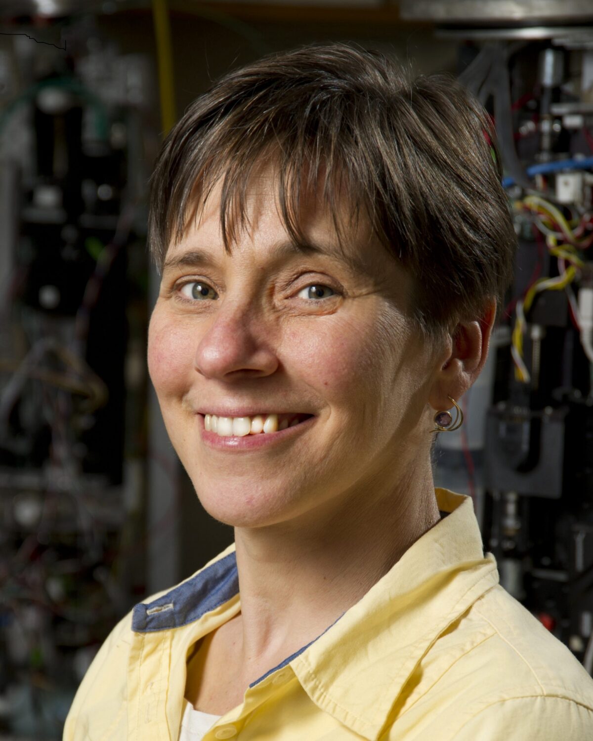 A person with short brown hair, wearing a yellow collared shirt, is smiling at the camera. The background shows blurred machinery and cables.