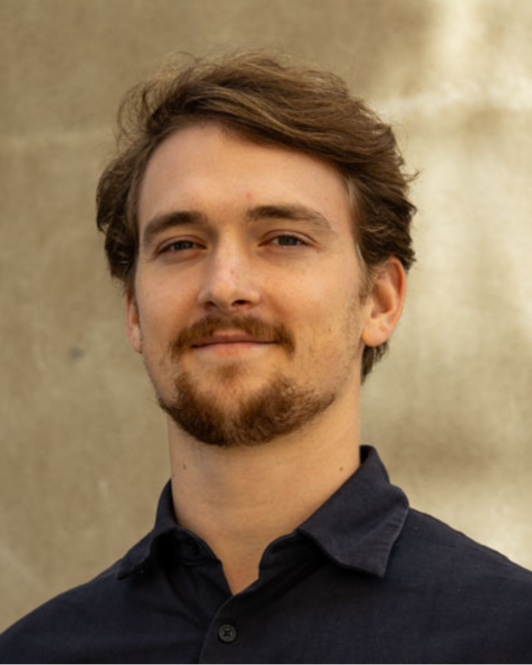 A man with light skin, short brown hair, and a beard is wearing a dark collared shirt and standing in front of a beige textured wall, looking at the camera with a slight smile.