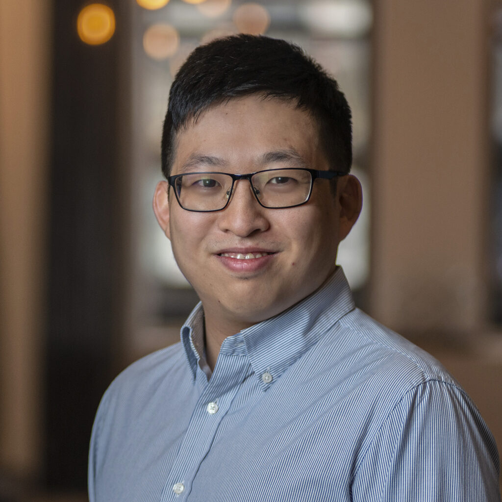 A person with short black hair and glasses, wearing a light blue button-up shirt, smiles at the camera in an indoor setting with blurred lights in the background.