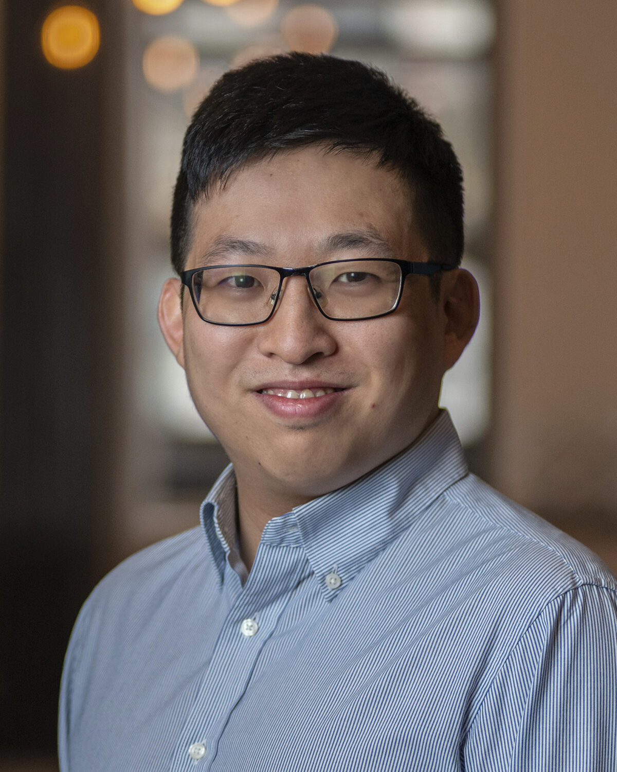 A person with short black hair and glasses, wearing a light blue button-up shirt, smiles at the camera in an indoor setting with blurred lights in the background.