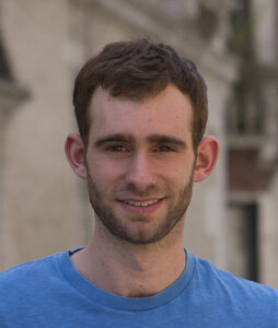 A young man with short brown hair and a trimmed beard wears a blue shirt and smiles at the camera. The background appears to be an outdoor setting with light stone walls.