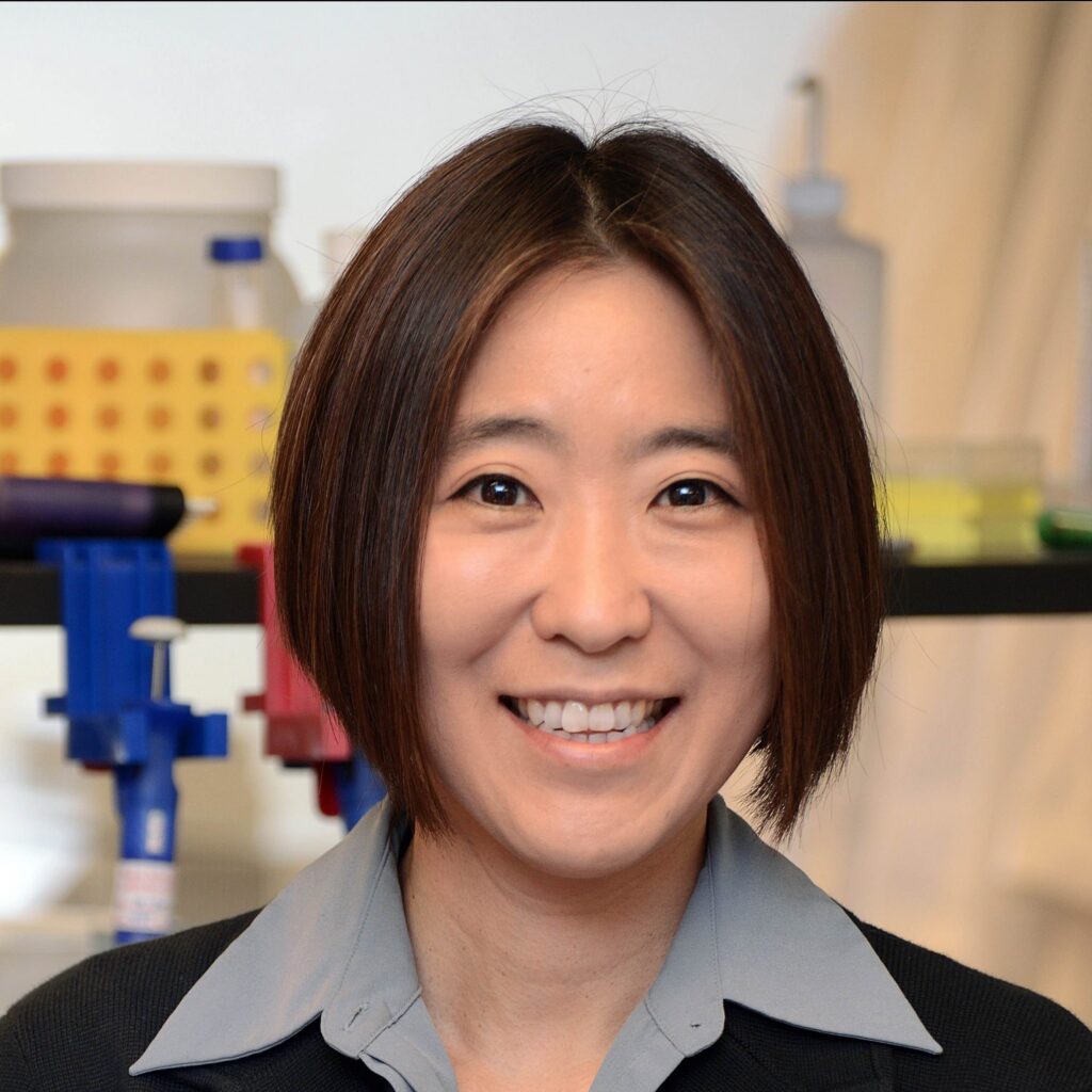 A person with short, straight hair smiles in a lab setting, with laboratory equipment and supplies visible in the background.