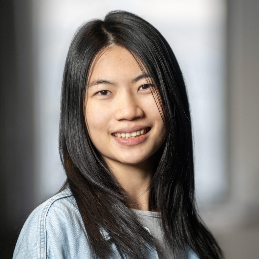 A young woman with straight black hair smiles at the camera, wearing a light denim jacket. The background is softly blurred, giving a warm and casual feel to the portrait.