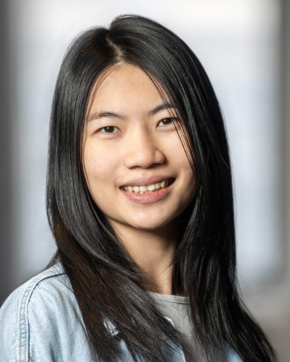 A young woman with straight black hair smiles at the camera, wearing a light denim jacket. The background is softly blurred, giving a warm and casual feel to the portrait.