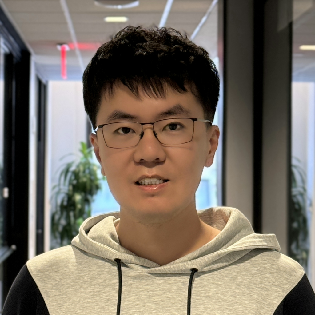 A young man wearing glasses and a light gray hoodie stands in a hallway with glass walls and potted plants in the background.