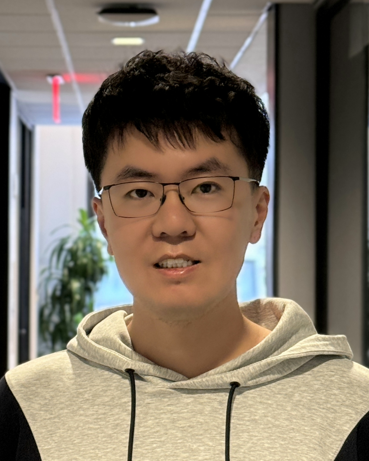 A young man wearing glasses and a light gray hoodie stands in a hallway with glass walls and potted plants in the background.