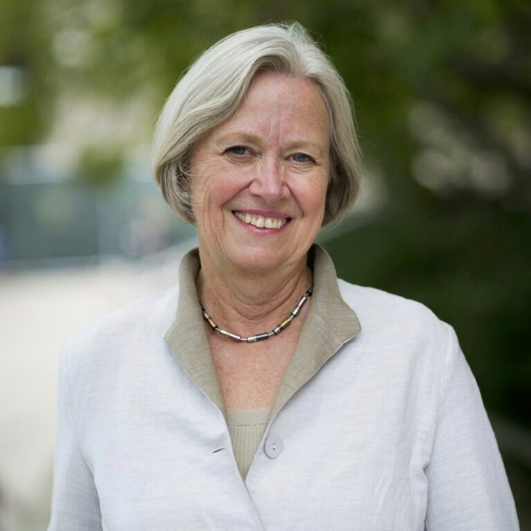 An older woman with short gray hair smiles at the camera, wearing a light-colored jacket and necklace, standing outdoors with greenery in the background.