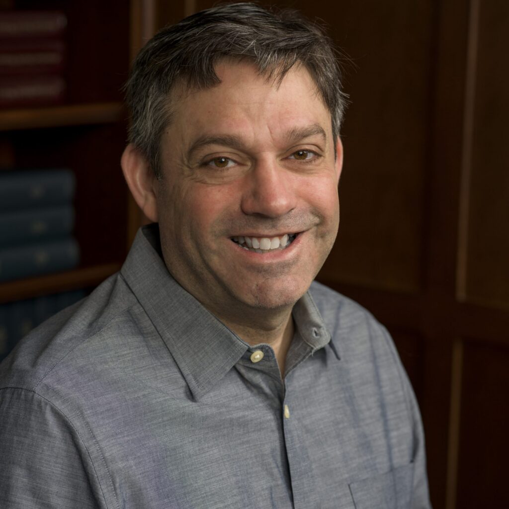 A man with short brown hair smiles at the camera, wearing a grey button-up shirt. He is seated in front of a wooden bookshelf filled with books.