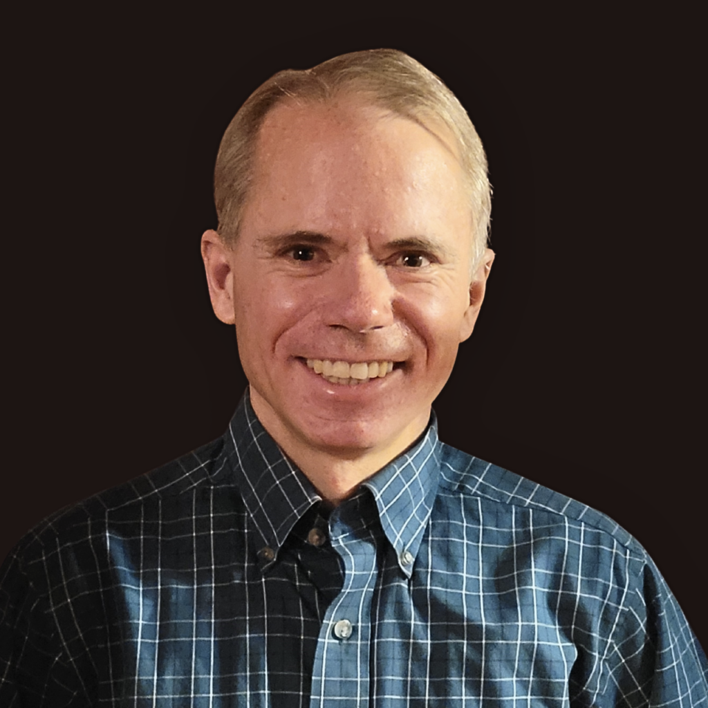 A smiling man with short light hair wearing a blue and white plaid button-up shirt, posed against a solid dark background.