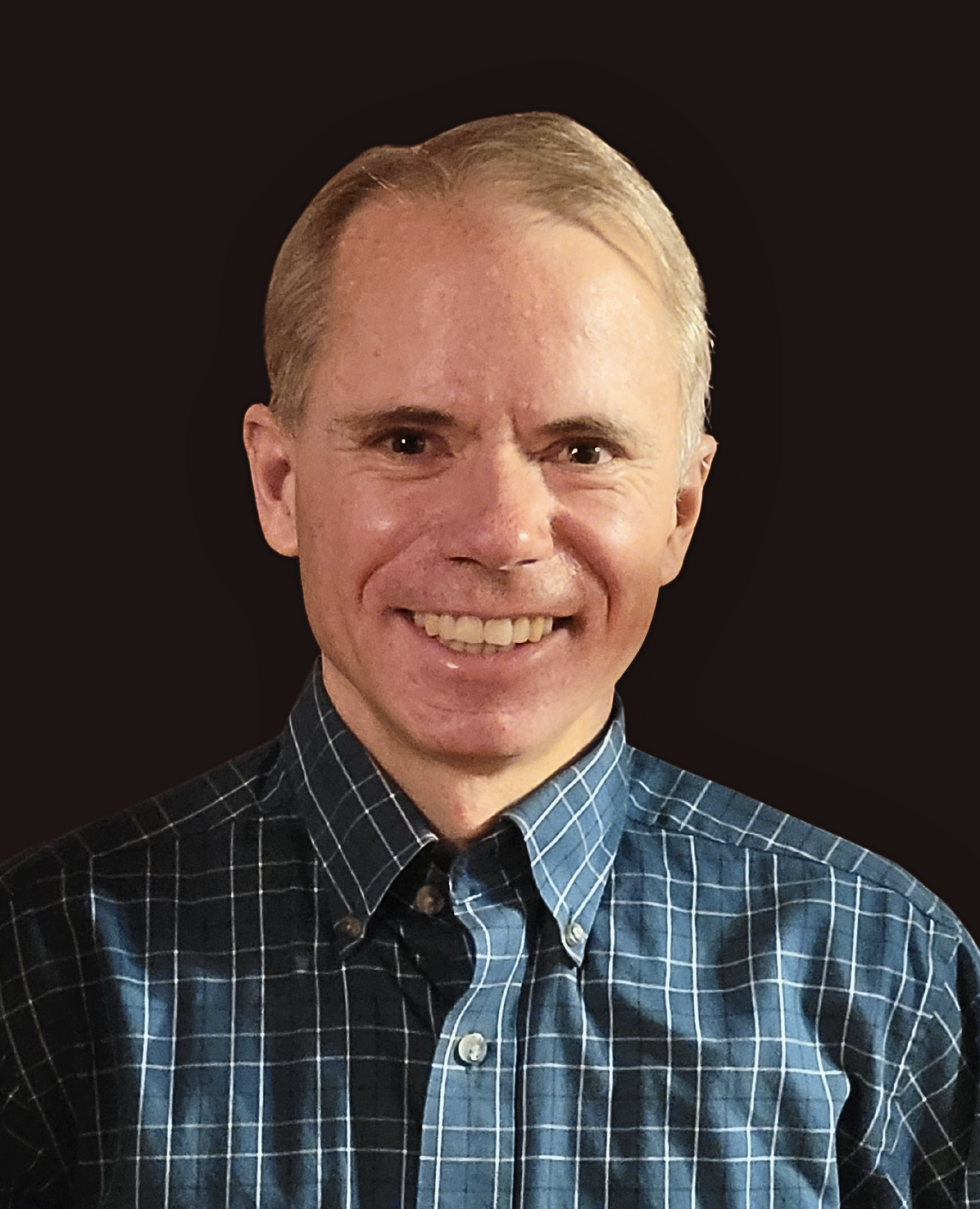 A smiling man with short light hair wearing a blue and white plaid button-up shirt, posed against a solid dark background.