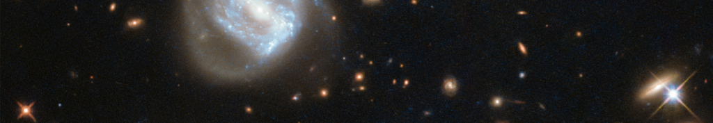 A spiral galaxy glows with blue-white light amid a dark space background, surrounded by numerous bright stars and distant galaxies.