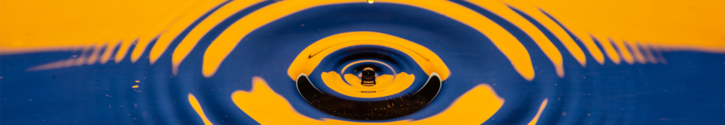 A photograph of what appears to be a drop of water breaking the surface of a pool of water and causing concentric circular ripples to form around the site of the impact.