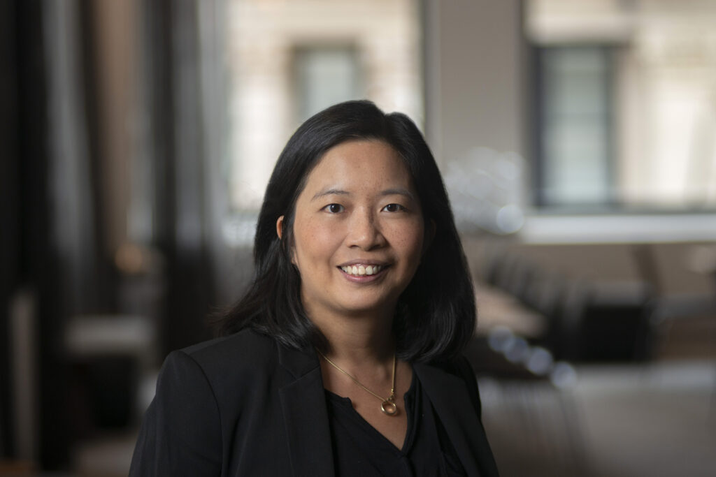 A woman with shoulder-length black hair, wearing a black blazer and gold necklace, smiles at the camera in a softly lit office setting with blurred windows in the background.