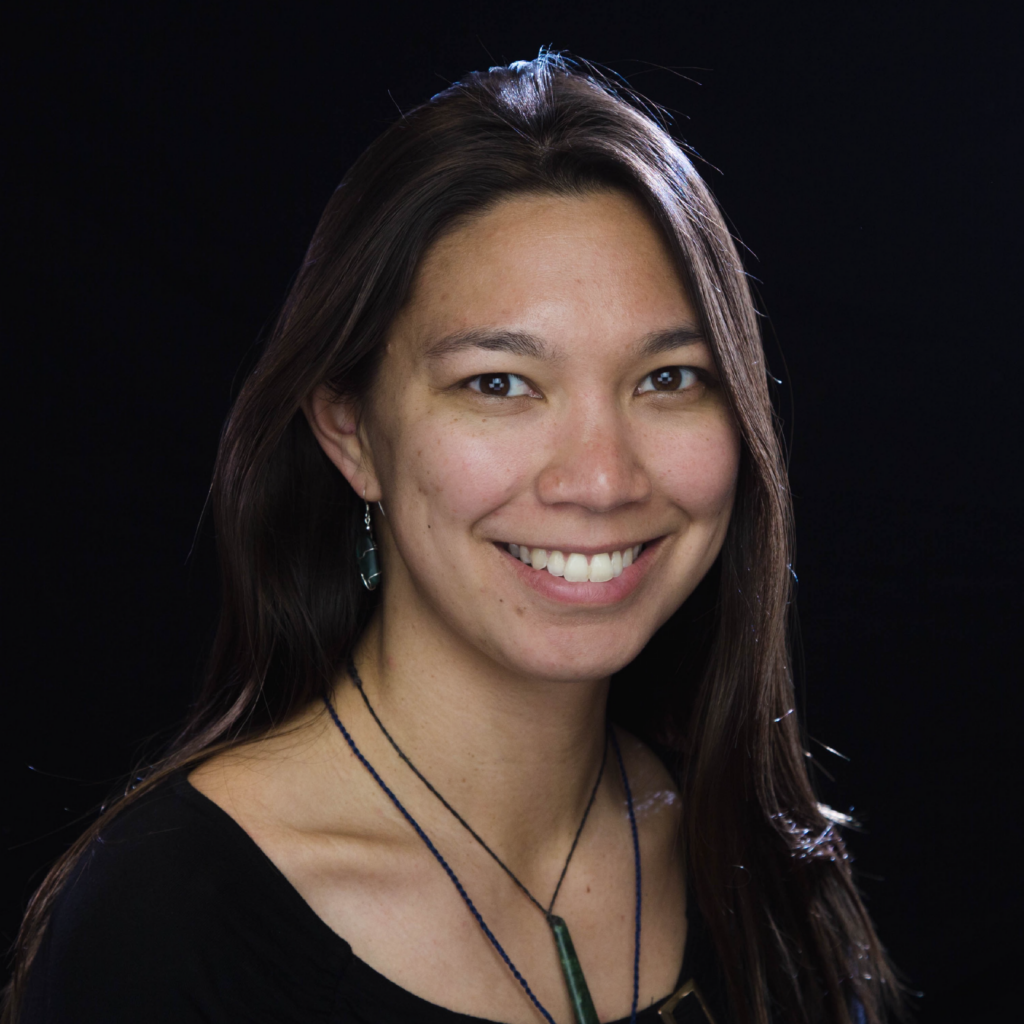 A woman with long brown hair, wearing a black top and necklaces, smiles at the camera against a black background.