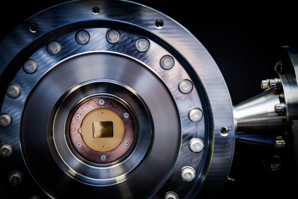 Close-up of a metallic scientific instrument with a circular design, bolts around the edge, and a small chip-like component in the center, likely part of a high-tech or quantum device.