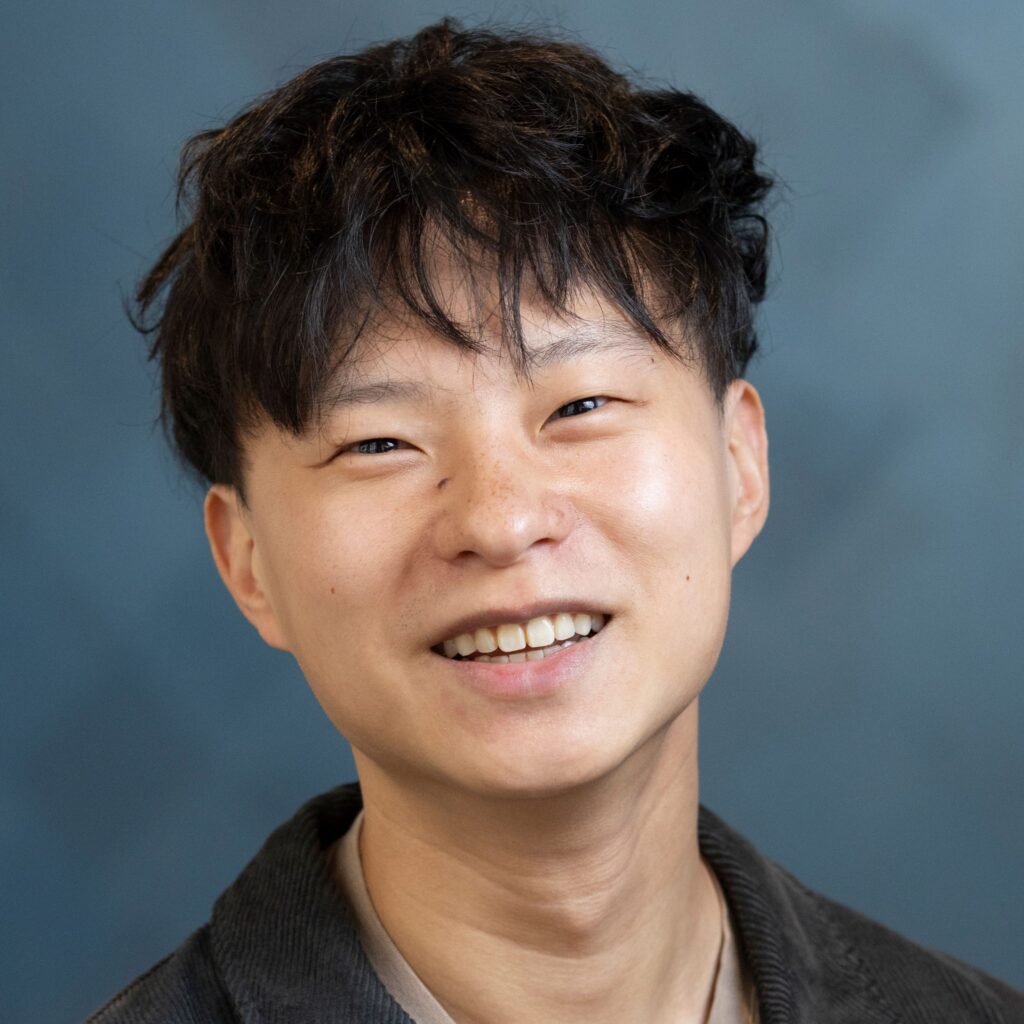 A young person with short, wavy black hair smiles warmly at the camera against a blurred blue background.
