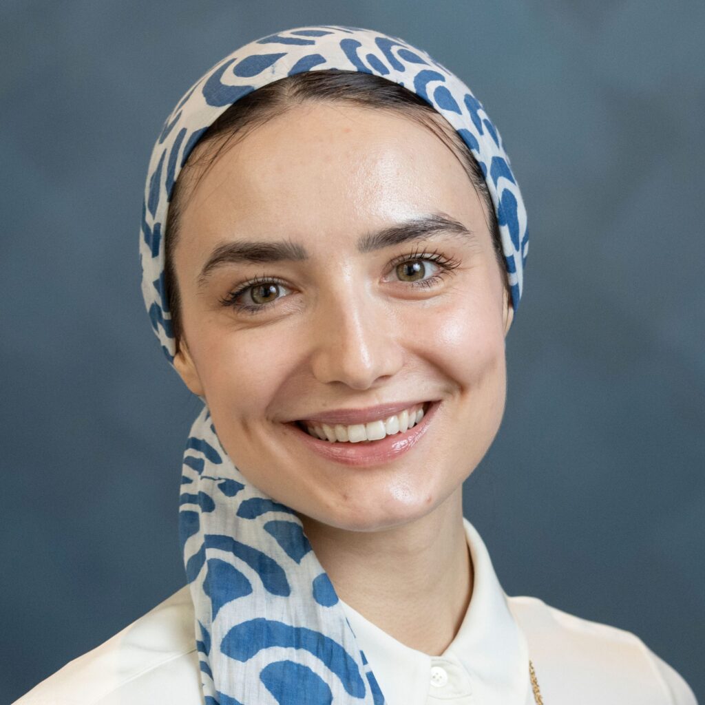 A person smiles at the camera, wearing a white shirt and a white headscarf with blue patterns. The background is a plain, dark blue.
