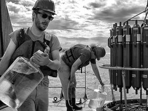 Two people on a boat collect water samples in large bottles, wearing safety vests and hard hats. Scientific equipment is visible, and the sky is cloudy.