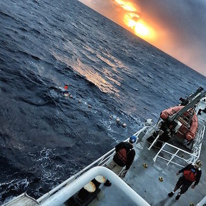 Two people in safety gear stand on a ships deck looking out at the ocean, with a bright orange sunset and dark clouds on the horizon.