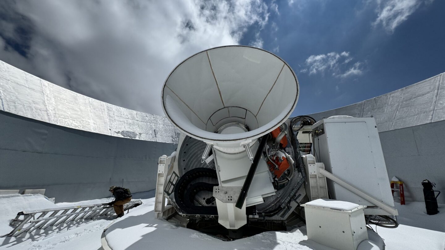 Large white satellite dish in a snowy observatory, surrounded by curved metallic walls under a partly cloudy sky. A ladder and various equipment are visible on the snow-covered ground.