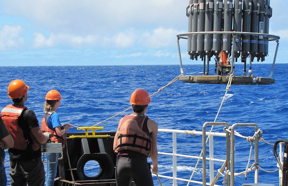 Three people wearing orange helmets and life vests stand on a ship, lowering a large scientific instrument into the blue ocean using ropes under a partly cloudy sky.